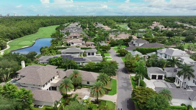 Residential neighborhood with luxury homes and lush greenery. Palm trees line the streets as the camera flies above modern properties. Golf greens and water features stretch in the distance. Aerial.