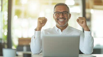 Obraz premium middle-aged Latin businessman with a broad smile, fists raised in celebration while reading great news on his smartphone, sitting at a modern office desk with a laptop open, embody