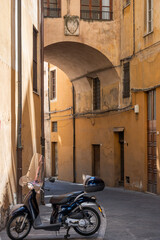 Iconic Italian lifestyle scene with scooter moped transport parked in overhead arched stone walled alleyway in streets of Pisa, Tuscany, Italy. 