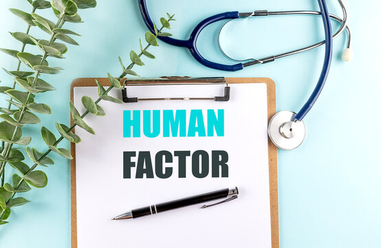 Clipboard with the text human factor on a light blue background, surrounded by a stethoscope, green leaves, and a pen