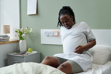 African American pregnant woman sitting on hospital bed holding her belly, showing emotion of discomfort, with flowers and medical equipment visible in room