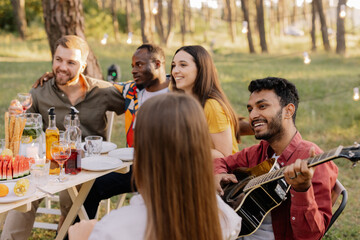 Multiracial group of people, Indian hipster man playing guitar and friends eating dinner and drinking wine during party in the forest