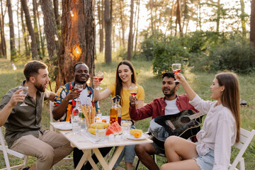 Meeting of multiracial group of friends playing guitar, singing, eating dinner and drinking wine during party in the forest