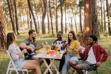Meeting of multiracial group of friends playing guitar, singing, eating dinner and drinking wine during party in the forest