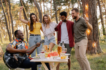 Multicultural group of people, African hipster man playing guitar and friends dancing, singing and having fun