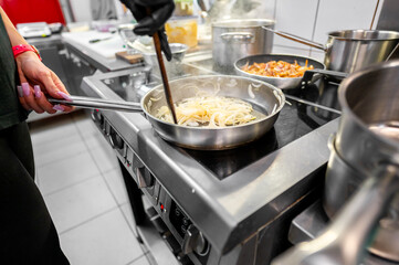 A chef’s hand stirs onion in a stainless steel pan on a gas stove, with various kitchen utensils and ingredients in the background. scene captures the essence of home cooking and culinary preparation.