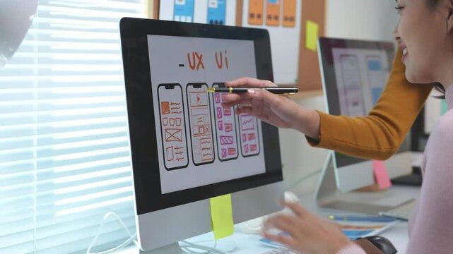Two female web designers are collaborating on a ux ui design project, using a desktop computer and pointing at the screen