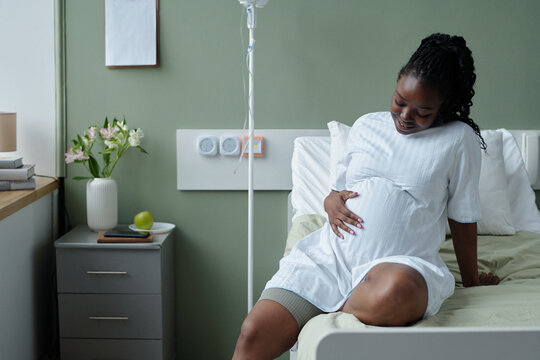 Smiling pregnant woman wearing a white gown sitting on a hospital bed, cradling her stomach and looking down thoughtfully. Table with flowers and medical equipment visible in background
