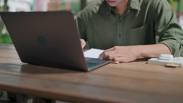 Close-up Asian man working seriously and taking notes while looking at laptop screen, outdoors in park during daytime, summer, book, pen and documents on table.