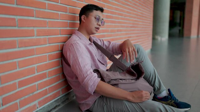 Asian man leaning against a brick wall holding a bag and looking up in a thoughtful manner and looking around, thinking about random things. Thinking about life, feeling sad and contemplative, absent-