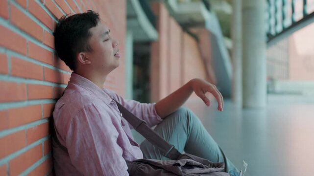 An Asian man is sitting with a very unhappy and disappointed look. He leans against a brick wall with his eyes closed and his hands on his head. He is feeling disappointed, sad, thinking, lost and dis