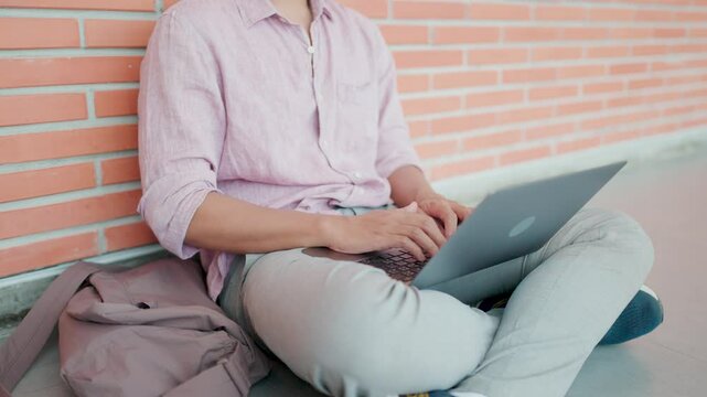 Close-up: Slow motion of Asian man's hand in pink shirt and glasses, sitting with bag next to him, leaning against brick wall on the floor and typing on laptop, freelancing, editing and working in rea