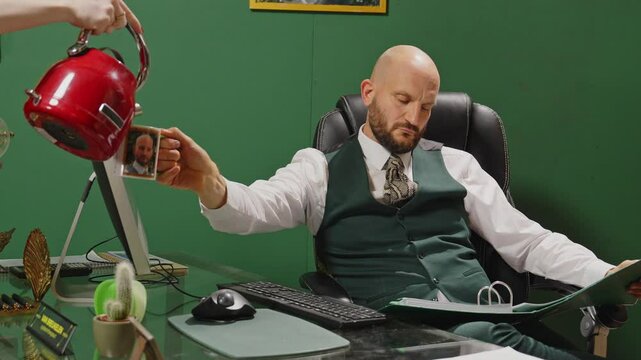 An arrogant manager holds out his cup while an assistent pours coffee in it. He talks to her and waves her away while reading a file