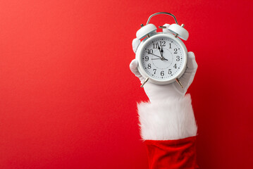 A Santa Claus hand holding a vintage alarm clock against a red background symbolizing holiday...