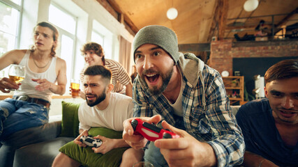 Cheers as two of guys playing football on console, laughter filling cozy room. Popcorn, chips and alcohol drinks keep group energized for their intense matches. Concept of football fans, sport evens