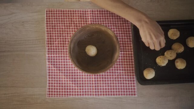 Person carefully places freshly baked chipa bread in a rustic wooden bowl on a red checkered cloth, showcasing south american culinary tradition of northeast riverside argentinian provinces