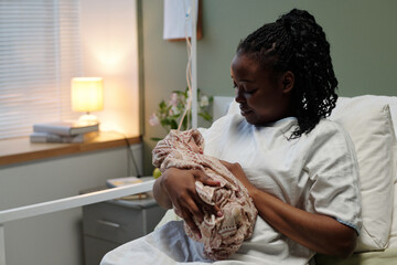 Black woman sitting in hospital bed in softly lit room, tenderly cradling her newborn while gazing...