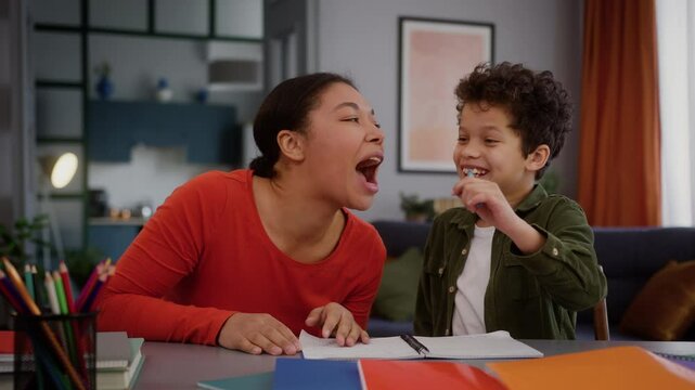 Happy African American family having fun funny play woman mom help with homework little son boy child kid together studying home education mother hold pen with lips pretend to have mustache laugh