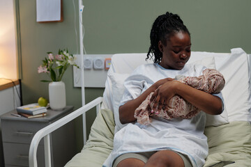 Caring mother holding newborn baby while sitting on hospital bed in maternity ward gently looking...