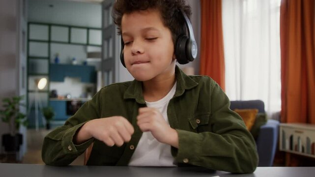 Funny African American son kid child boy pupil at home having fun positive pause break listening music in headphones listen audio song dancing with hands moving enjoy dance holding mobile phone move
