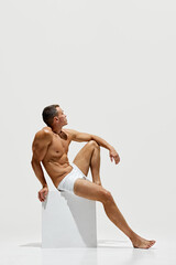Fototapeta premium Like a statue. Handsome man posing shirtless on cube, showing his perfect muscular body isolated on white studio background. Concept of male beauty, body and skin care, health, sport