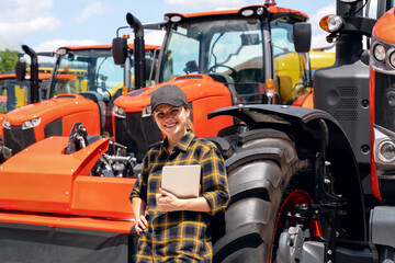 Agricultural or Construction equipment dealer. Woman in plaid shirt and cap with digital tablet in her hand leaned on the wheel of the new tractor.