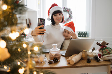Caucasian mother and young child wear Santa hats taking selfie in cozy kitchen setting. Baking supplies and Christmas decor suggest festive holiday celebration.