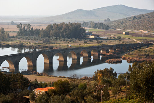 An ancient stone bridge spanning across a river, with a picturesque rural landscape featuring gentle hills, trees, and fields in the background, on a clear day in Medellin Andalusia Spain