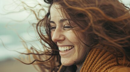A lively and joyful woman with wild, windswept hair laughs heartily at the beach on a blustery day.