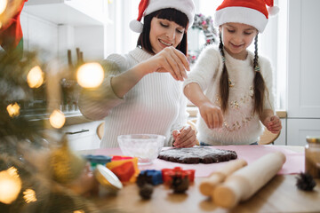 Caucasian mother and daughter wearing Santa hats sprinkle flour on dough in festive kitchen. Joyful holiday atmosphere captured while baking Christmas cookies.