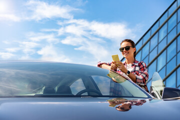 A female car driver stands next to her new vehicle, using her smartphone in front of a business centre.