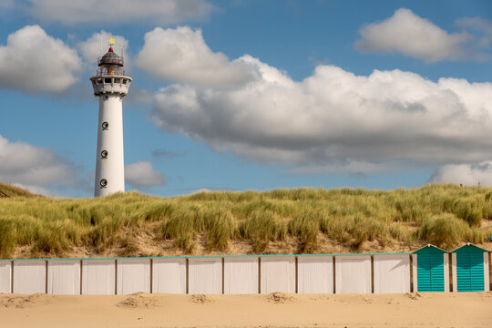The tall lighthouse of Egmond aan Zee, Netherlands, beside sandy dunes and beach huts under a bright blue sky with fluffy clouds on a sunny day