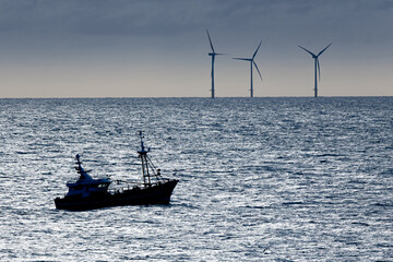 Silhouette of a fishing boat navigating the ocean near wind turbines at dusk with dark clouds...
