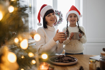 Smiling Caucasian mother and young daughter drinking hot cocoa in cozy kitchen. Both wearing Santa...