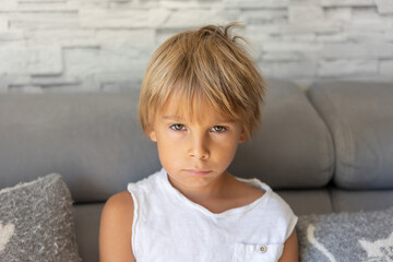 Blond child, cute preschool boy with swollen face from a toothache, sitting on a couch at home in...