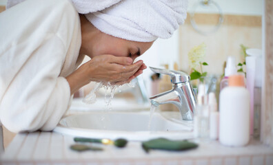 A woman in a towel and bathrobe washes her face at a bathroom sink with water splashing from her hands, surrounded by skincare products, symbolizing a morning routine, self-care, and cleanliness.