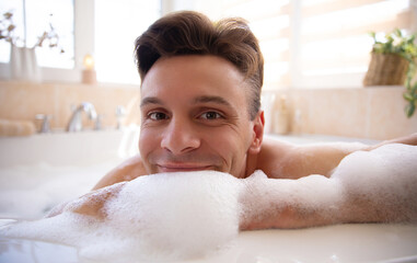 A cheerful man relaxes in a bubble bath, resting his chin on the edge, surrounded by foam. The bright bathroom with soft lighting and minimal decor creates a serene and cozy atmosphere.