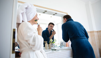 A man in a blue bathrobe shaves in front of a bathroom mirror while a woman in a white bathrobe, with a towel on her head, uses a face roller. The couple enjoys a shared morning skincare routine.