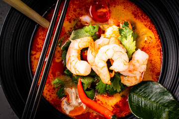 Spicy Thai Tom Yum Kung soup with shrimp, lemongrass and galangal, cilantro, cherry tomatoes and mushrooms in a black bowl on a graphite background with chopsticks and a spoon. Close-up.