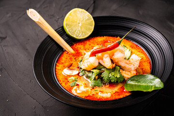 Spicy Thai Tom Yum Kung soup with shrimp, lemongrass and galangal, lime and chili pepper, cilantro, cherry tomatoes and mushrooms in a black bowl on a graphite background