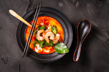 Spicy Thai Tom Yum Kung soup with shrimp, lemongrass and galangal, cilantro, cherry tomatoes and mushrooms in a black bowl on a graphite background with chopsticks and spoon