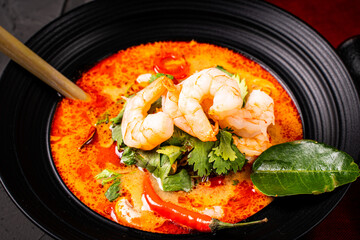 Spicy Thai Tom Yum Kung soup with shrimp, lemongrass and galangal, cilantro, cherry tomatoes and mushrooms in a black bowl on a graphite background with chopsticks and a spoon. Close-up.