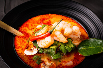 Spicy Thai Tom Yum Kung soup with shrimp, lemongrass and galangal, cilantro, cherry tomatoes and mushrooms in a black bowl on a graphite background with chopsticks and a spoon. Close-up.