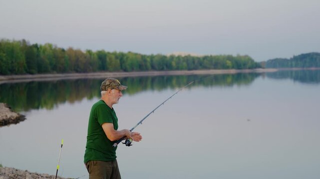 Portrait shot of a senior angler casting fishing rod along the river coast at dusk. Active adult fishing in river.