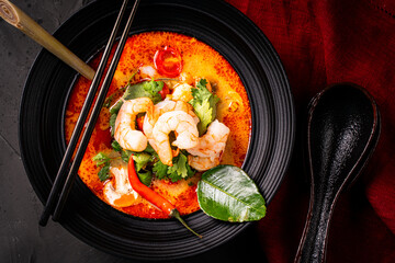 Spicy Thai Tom Yum Kung soup with shrimp, lemongrass and galangal, lime and chili pepper, cilantro and mushrooms in a black bowl on a dark background with chopsticks and a spoon and a burgundy napkin.