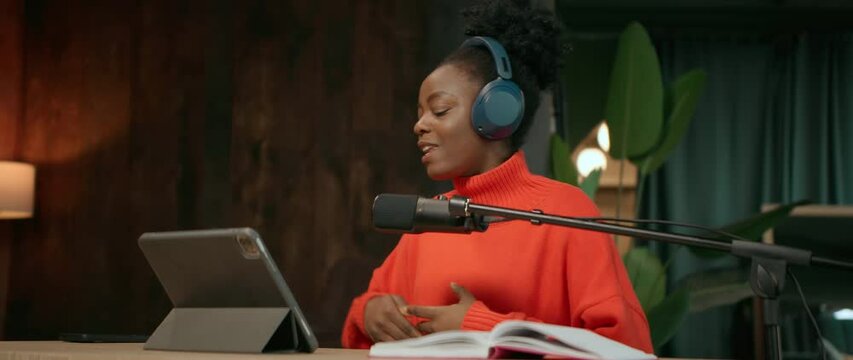 Footage of energetic attractive young African American woman with stunning afro hairstyle talking into microphone while recording radio show in modern studio. Radio host using digital tablet. Camera