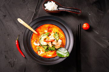 Tom yam kung, spicy Thai soup with shrimp, lemongrass and galangal, lime, chili, cilantro in a black bowl on a dark background with chopsticks and a spoon with rice.