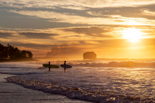 Two surfers holding surfboards, looking at the waves against a vibrant sunrise at Playa El Tunco, a popular surf beach destination in El Salvador. Sunshine shines over the ocean and iconic rock stack.