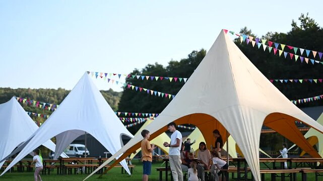 Families and friends gather and socialize at an outdoor festival event. White and tan peaked tents are set up with colorful flags strung between them. Picnic tables are on the lawn around the tents.