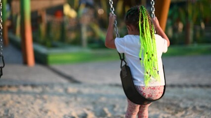 Little girl with green braids playing on a swing set. She swings back and forth by herself on a playground.